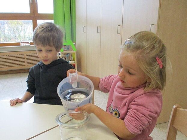 Zwei kleine Kinder, ein Mädchen und ein Junge an einem Tisch. Das Mädchen gießt Wasser aus einem Messbecher in ein Glasgefäß.