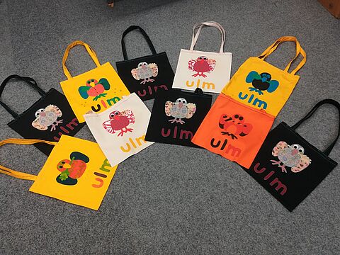 Stofftaschen in verschiedenen Farben mit aufgenähtem Ulm-und Spatzenemblem.